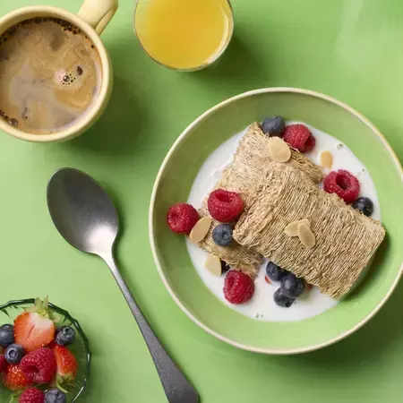 Shredded wheat, milk and berries in a green bowl with a coffee and orange juice alongside.