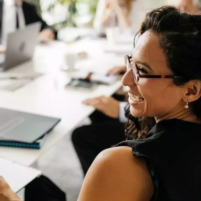Woman smiling while sitting at a work table with colleagues in the background.